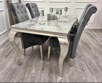 A grey marble dining table with a silver metal frame accompanied by four grey velvet chairs with chrome lion knocker backrest details.